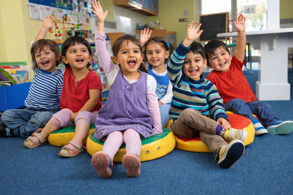 Happy preschool children sitting in a classroom, raising their hands and smiling during a group learning activity.